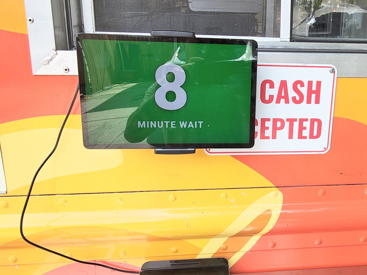 A tablet mounted in a food truck's service window showing a green screen with a large 8 and the words MINUTE WAIT